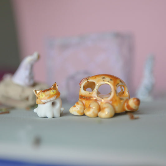Two miniature ceramic figurines, including a white cat with an orange-tan face and a small orange-tan shell-like creature, displayed on a tabletop with a soft pink blurred background.